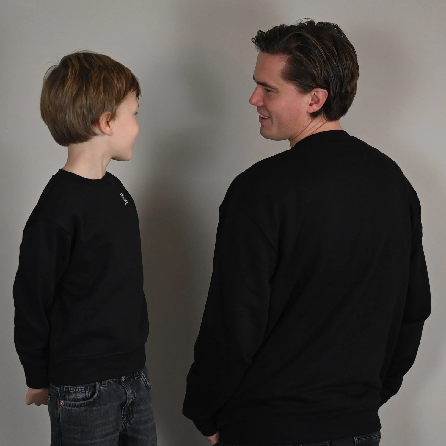 vader en zoon dragen dezelfde toebie sweater in het zwart terwijl ze elkaar aankijken en lachen