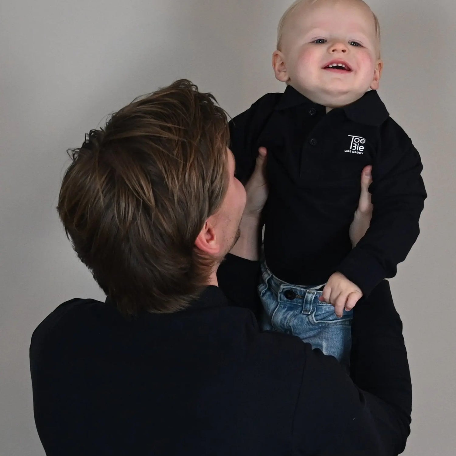 vader en zoon dragen dezelfde toebie polo met lange mouw, vader tilt zijn zoon op terwijl het zoontje lacht
