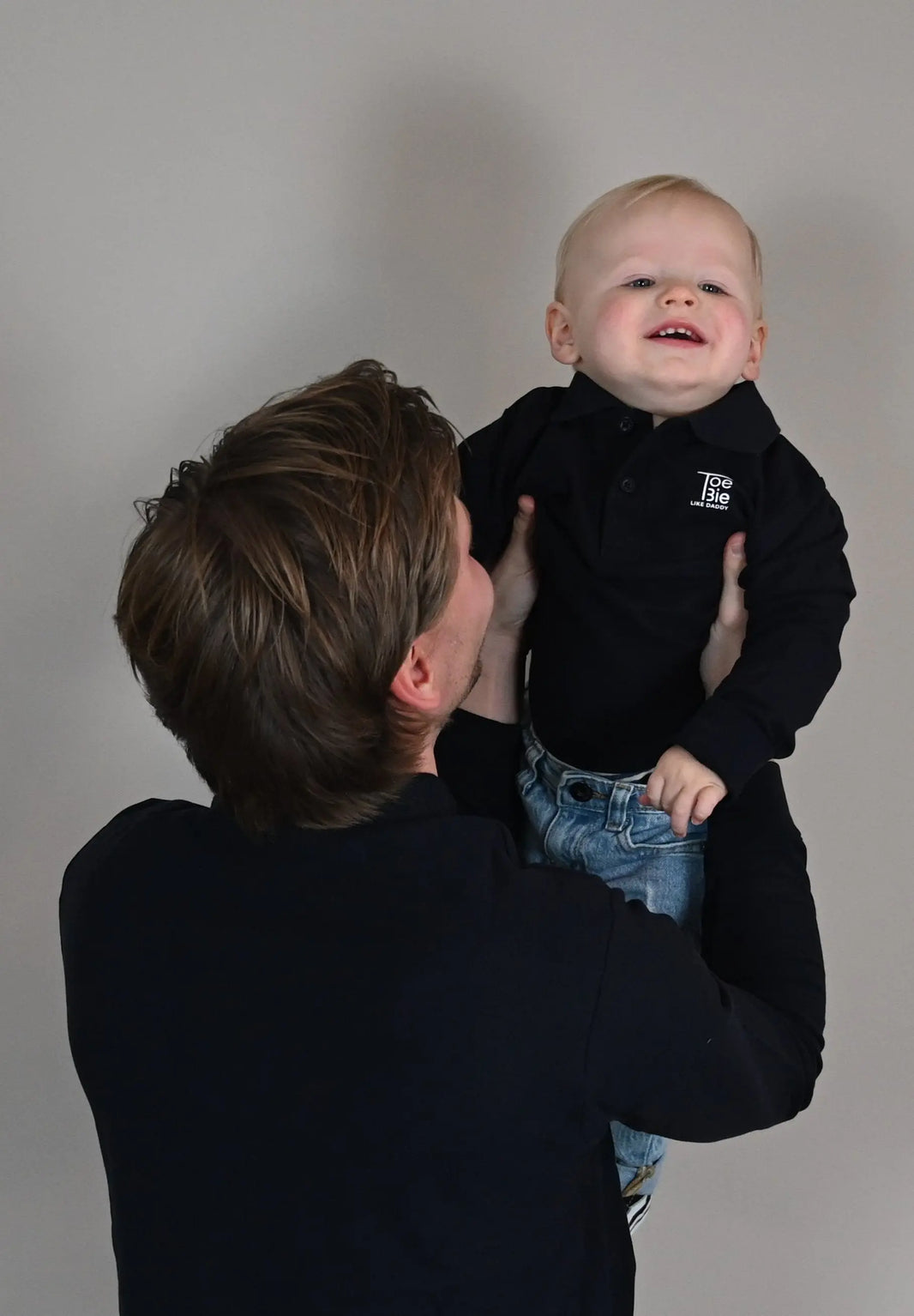 vader en zoon dragen dezelfde toebie polo met lange mouw, vader tilt zijn zoon op terwijl het zoontje lacht