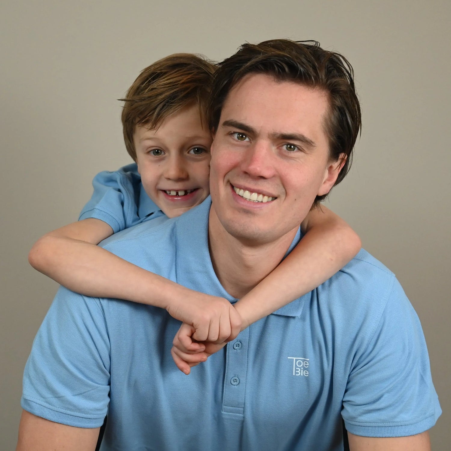 Vader en zoon dragen dezelfde outfit en kijken lachend in de camera terwijl de zoon op zijn vader zijn rug zit. 